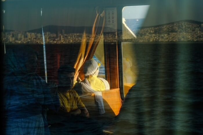 Street photo capturing everyday life inside a ferry with passengers reflected against a cityscape and water backdrop.