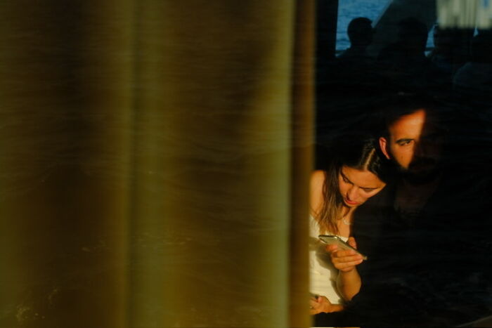 Shadowed street photo of a woman and man capturing everyday life with a smartphone detail in natural light.