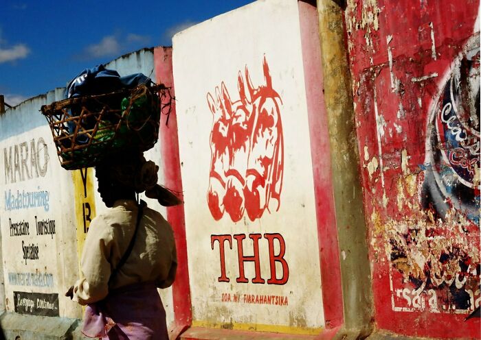 A street photo showing a person carrying a basket on their head beside colorful painted walls in everyday life.