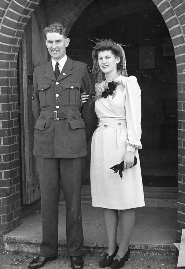 Bride in 1940s dress with groom in military uniform standing outside a brick building in a moving wedding photo.