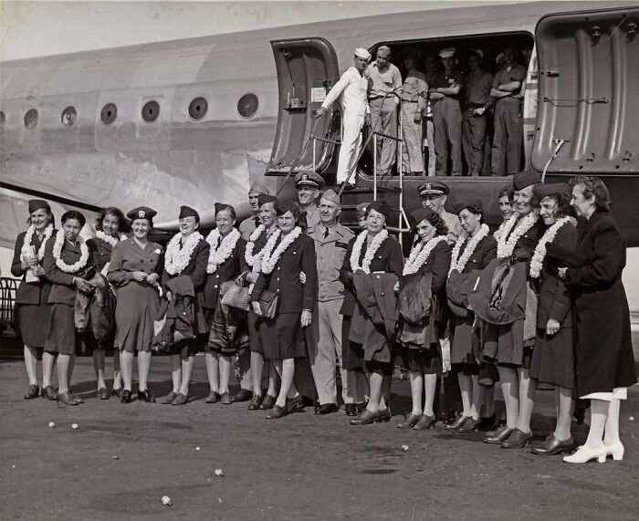 Group of women and military personnel wearing leis in front of WWII airplane, captured in rare WWII photo.