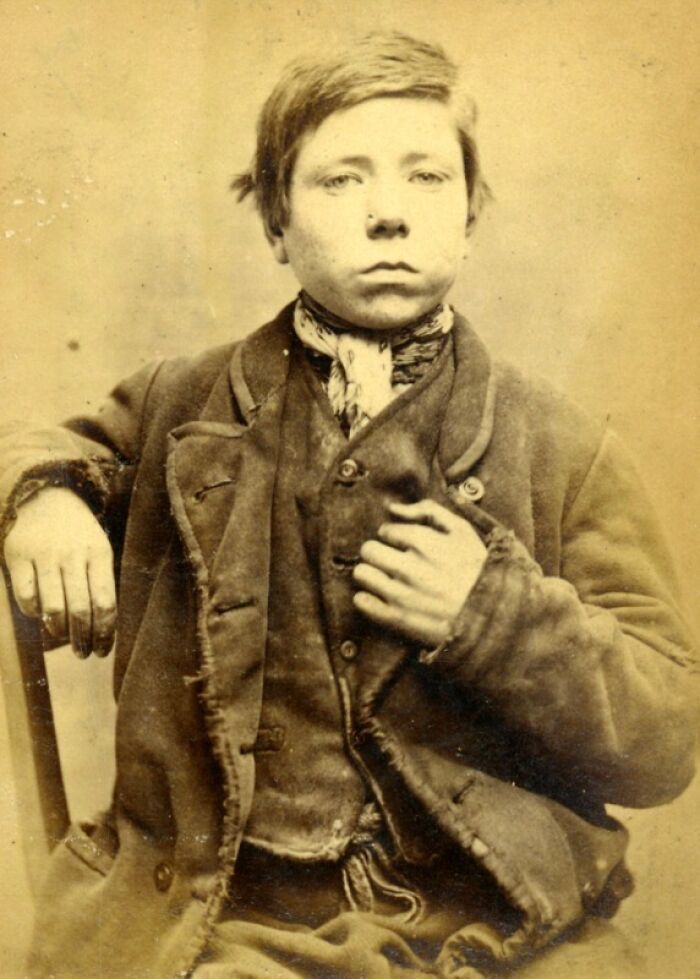 Vintage historical mugshot of a young boy in worn clothing seated and looking directly at the camera.