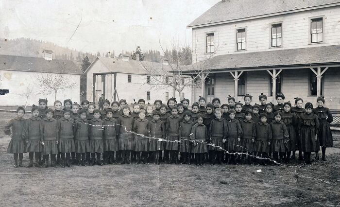 Group of Native American children lined up outside a boarding school building in an early 1900s haunting photo.