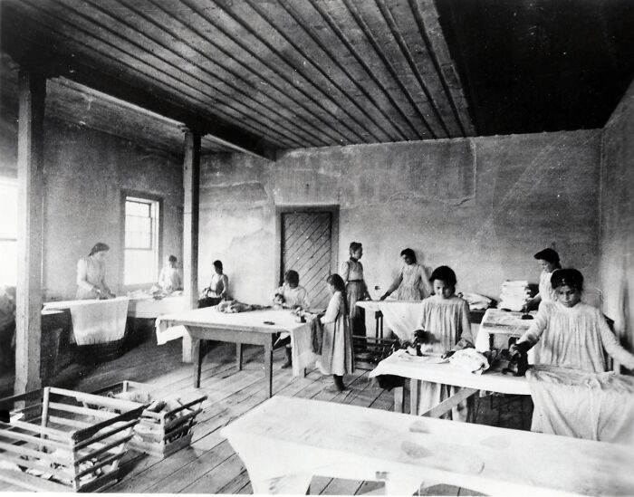 Native American boarding school students in the 1910s engaged in chores inside a sparse classroom setting.