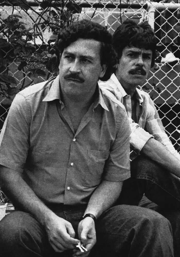 Black and white photo of two men sitting by a chain-link fence, representing notorious mob bosses in history.