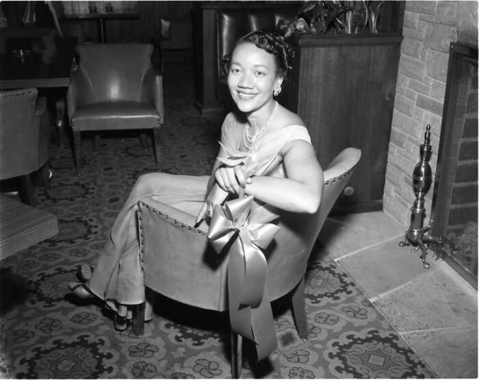 African American woman in 1960s dress sitting on a chair with a decorative bow, enjoying life in Los Angeles.