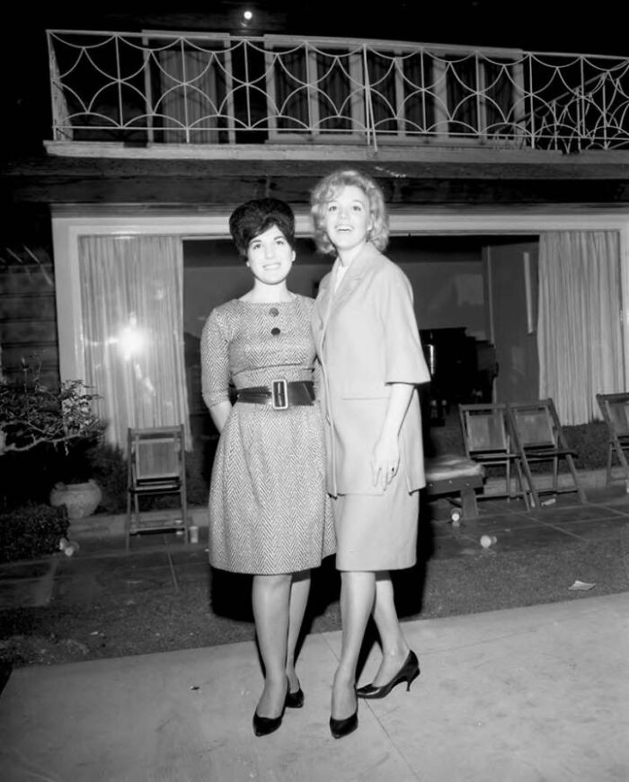 Two LA women dressed in 1962 fashion standing outside a home, enjoying a social moment in vintage style.