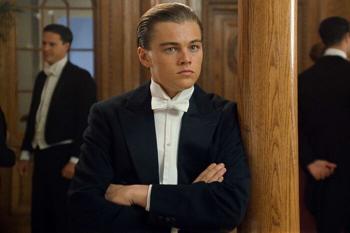 Young male actor in a black tuxedo and white bow tie posing confidently among others in a vintage setting, iconic heartthrob photo.