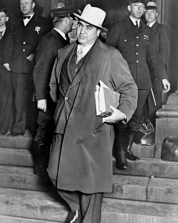 Notorious mob boss in a suit and hat walking down steps with officers behind, symbolizing infamous mob bosses history.
