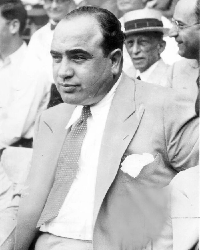 Black and white photo of a notorious mob boss in a suit and tie, seated outdoors among other men in vintage attire.