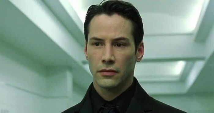 Keanu Reeves in a black suit standing in a futuristic hallway, a classic photo of iconic heartthrob actors.
