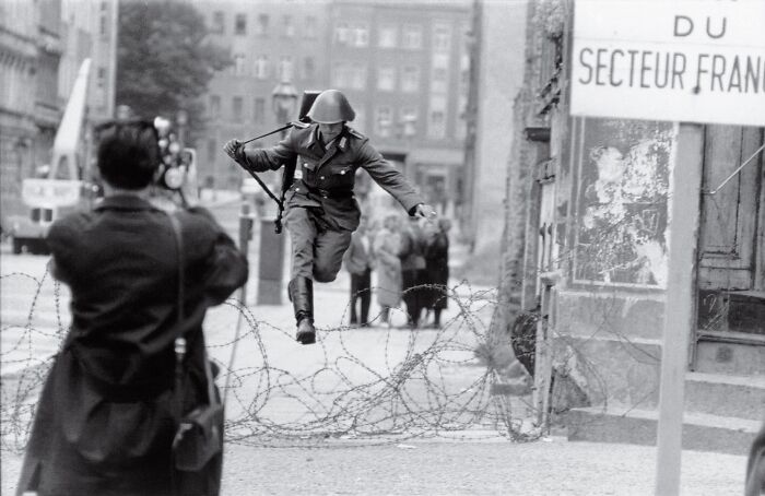 Soldier leaping over barbed wire barrier during a tense moment in history, captured in a striking black and white photo.