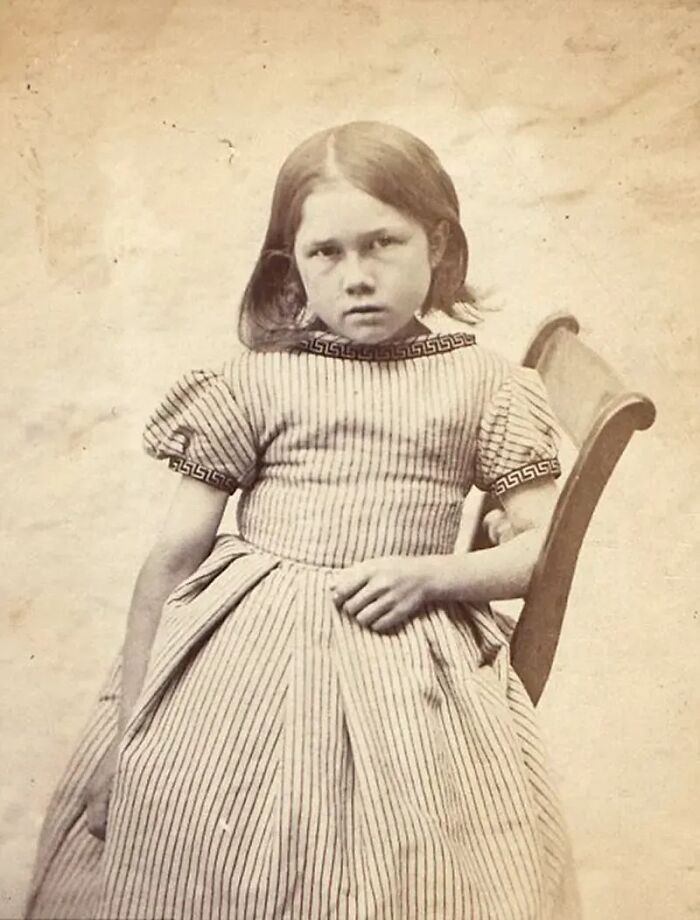 Vintage historical mugshot of a young girl in a striped dress sitting on a chair with a serious expression.