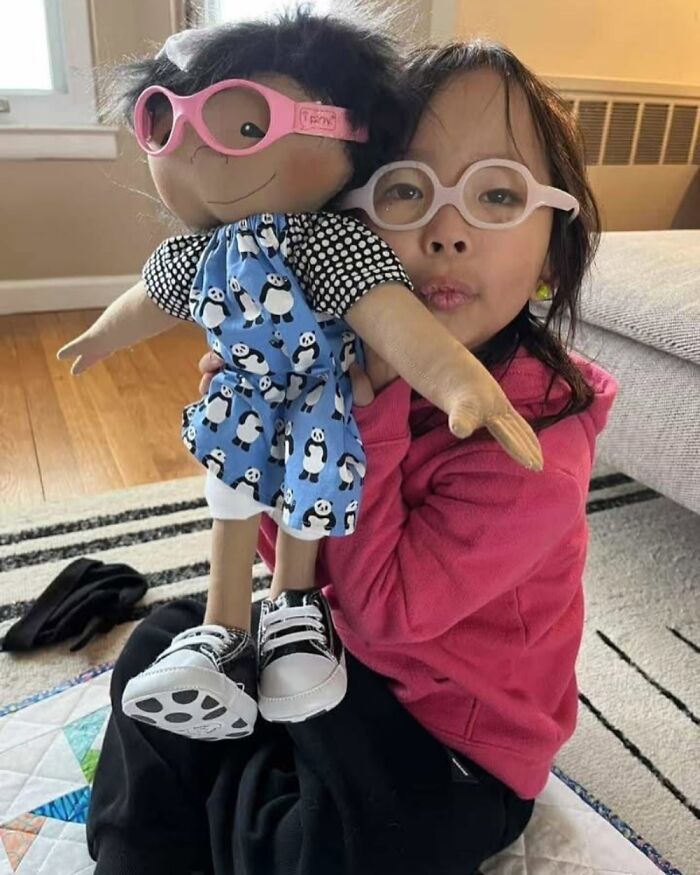 Child holding a custom doll designed to represent rare health conditions, both wearing glasses and smiling indoors.