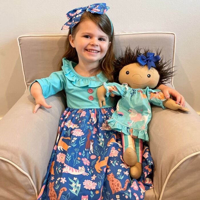 Young girl smiling while holding a custom doll representing her rare health condition in matching outfit and bow.