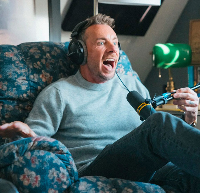 Dax Shepard wearing headphones, speaking into a microphone, reacting excitedly during a podcast recording session.