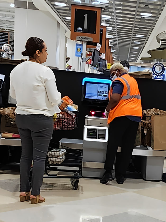 Customer with a cart waiting while cashier in orange vest uses self-checkout, illustrating selfish people making life harder for others.