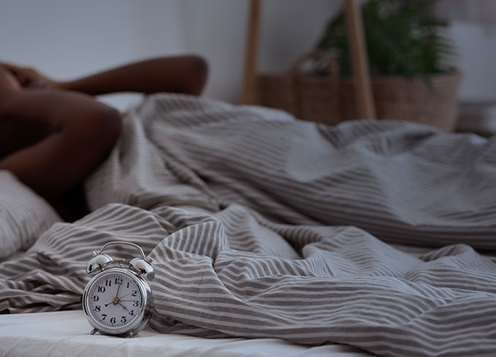 Alarm clock on bed with striped sheets and a man lying awake, highlighting a rare medical mystery of not sleeping for two years.