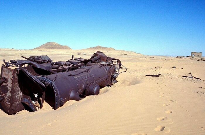 Rusting old train wreck half buried in desert sand, revealing unfamiliar parts of history in a harsh landscape.