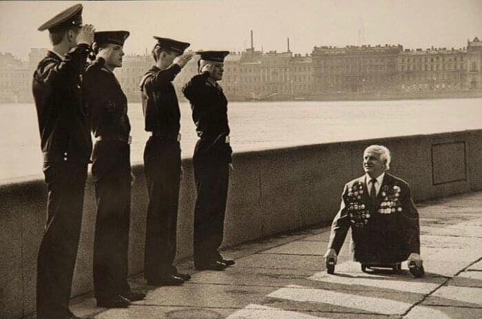 Four uniformed men salute a decorated war veteran with no legs on a historic waterfront, showcasing unfamiliar parts of history.