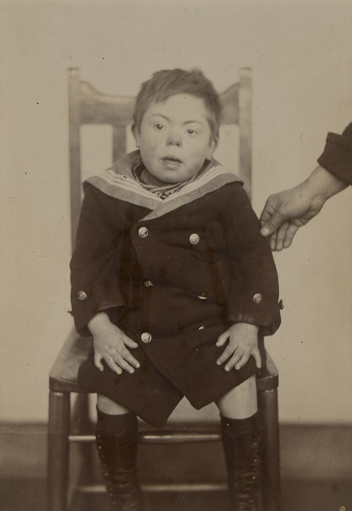 Black and white vintage photo of a young child seated on a wooden chair showing unfamiliar parts of history.