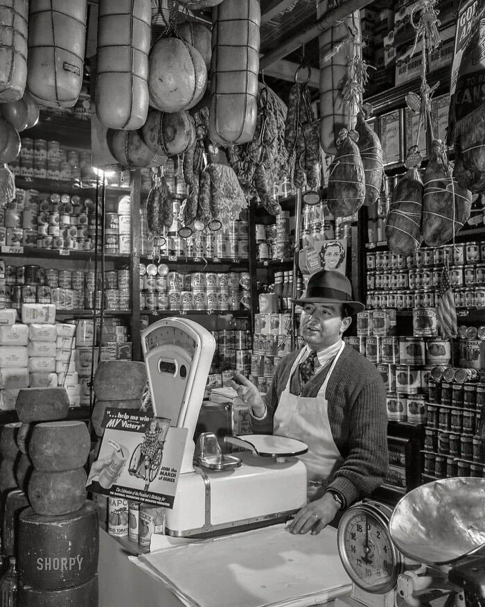 Man in vintage store with hanging meats and canned goods, showcasing unfamiliar parts of history in a black and white photo.