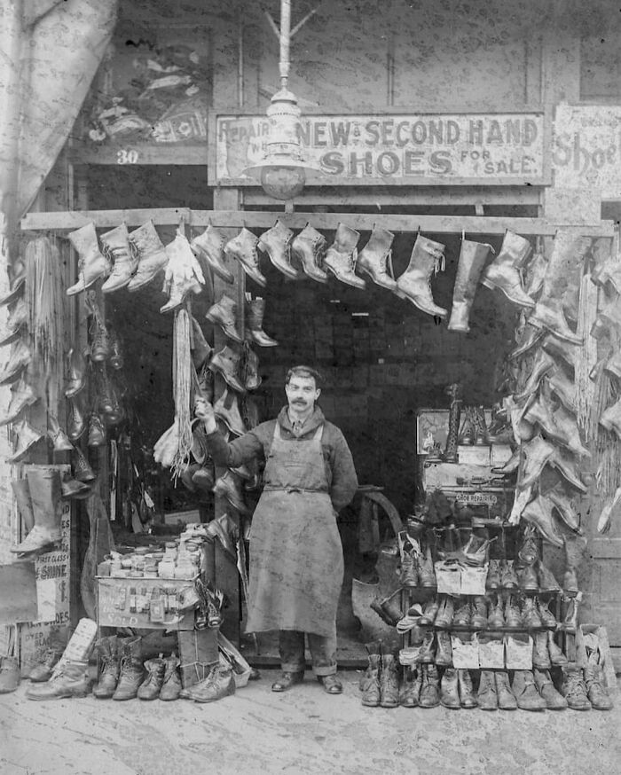 Historic black and white photo of a shoemaker in his workshop with boots for sale, showcasing unfamiliar parts of history.