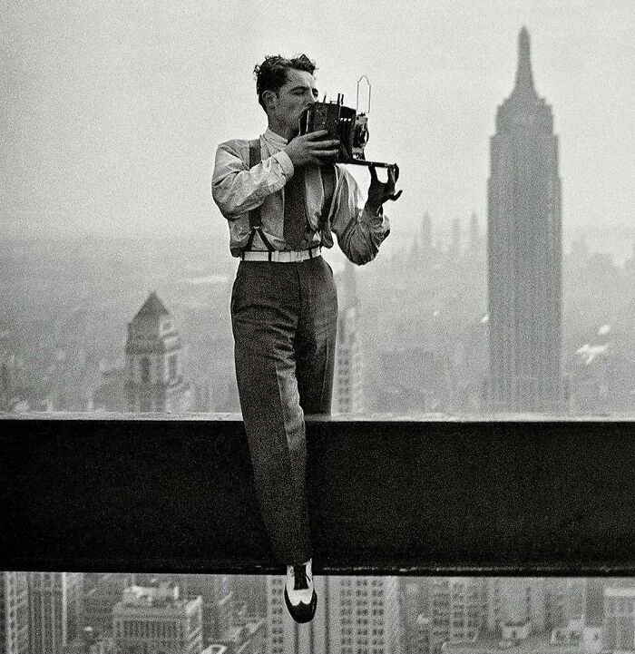 Man perched on a steel beam high above city skyline, capturing rare historical moments with vintage camera for unfamiliar parts of history
