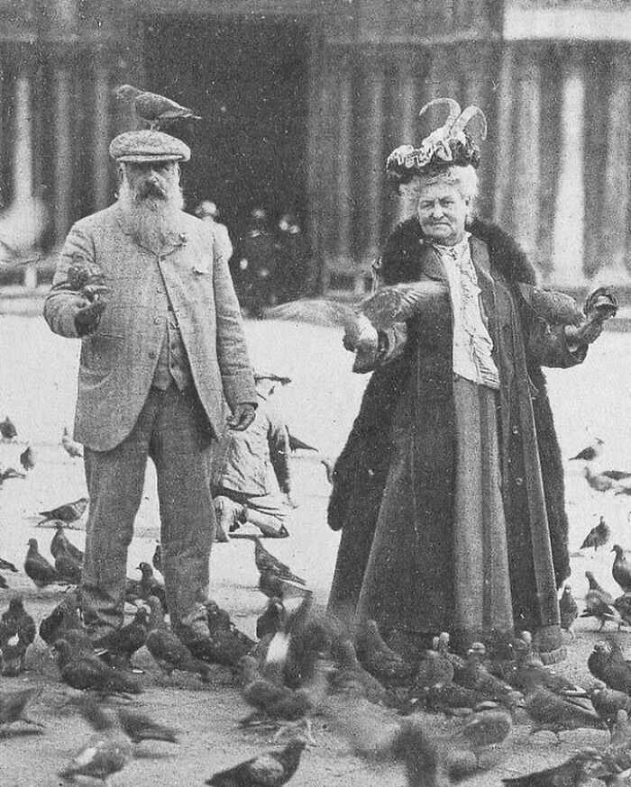 Black and white historical photo of an elderly couple surrounded by pigeons, showing unfamiliar parts of history.