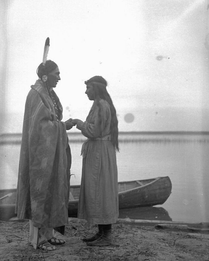Two Native Americans in traditional clothing standing by a lake with a canoe, revealing unfamiliar parts of history.