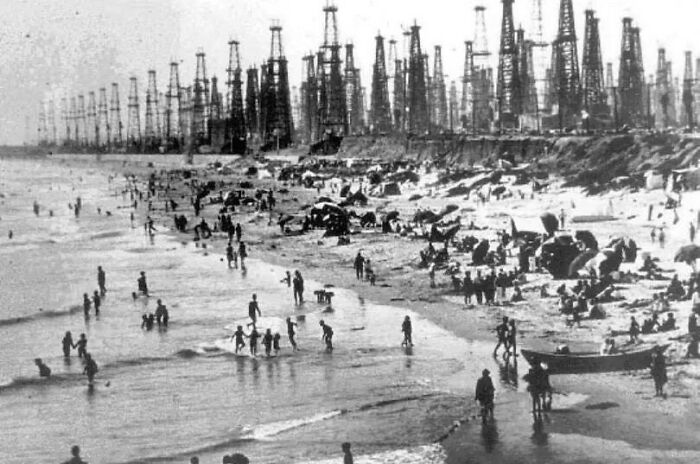 Crowded beach with numerous oil rigs in the background, showcasing unfamiliar parts of history through old industrial development.