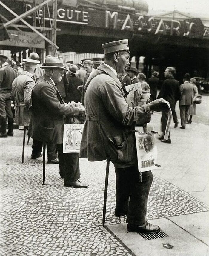 Men with amputated legs selling newspapers on a city street, showcasing unfamiliar parts of history through lived experiences.