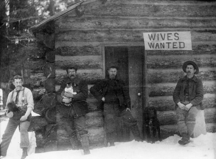 Four men in front of a log cabin with a "Wives Wanted" sign highlighting unfamiliar parts of history.