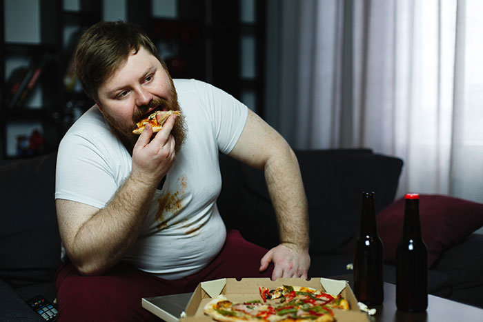 Man eating pizza on couch, enjoying new facts while relaxing with beer bottles nearby in a casual setting.