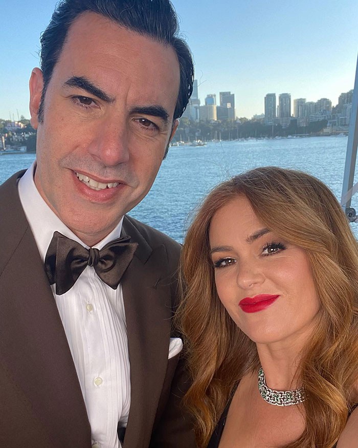 Isla Fisher and Sasha Baron Cohen smiling together outdoors with city skyline and water in the background. - 15
