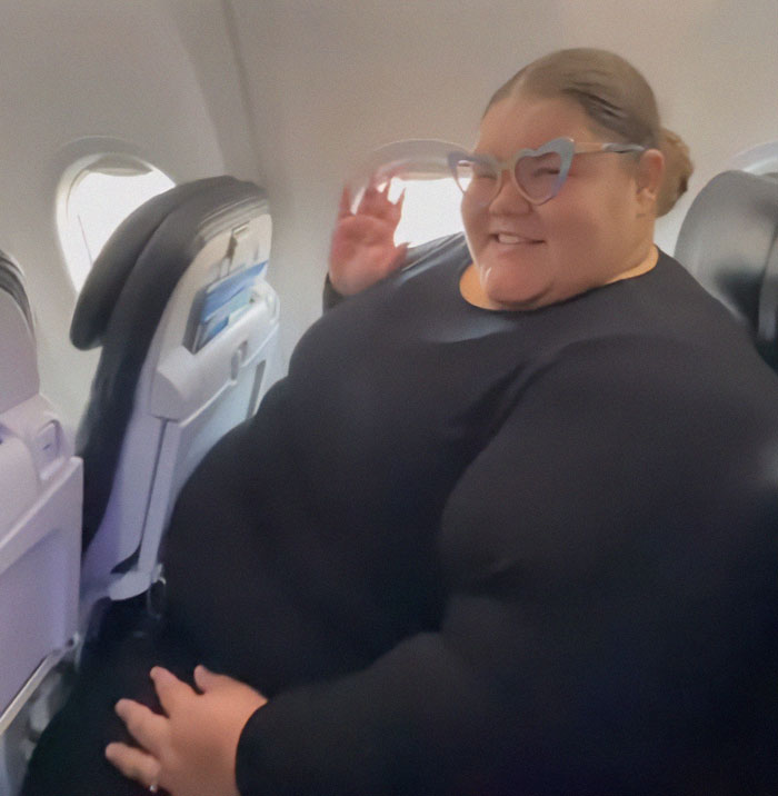 Plus-size passenger seated on an airplane, smiling and wearing heart-shaped glasses amid airline controversial new rule discussion. Plus-size passenger seated on an airplane, smiling and wearing heart-shaped glasses amid airline controversial new rule discussion.