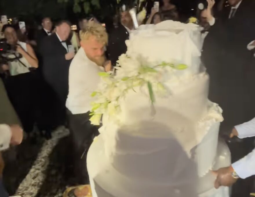 Jake Paul smashing a large wedding cake at a crowded event with people taking photos in the background
