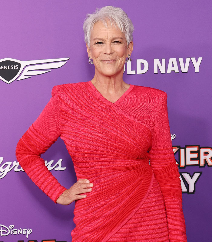 Jamie Lee Curtis posing on the red carpet, wearing a vibrant red textured dress and smiling confidently. - 1
