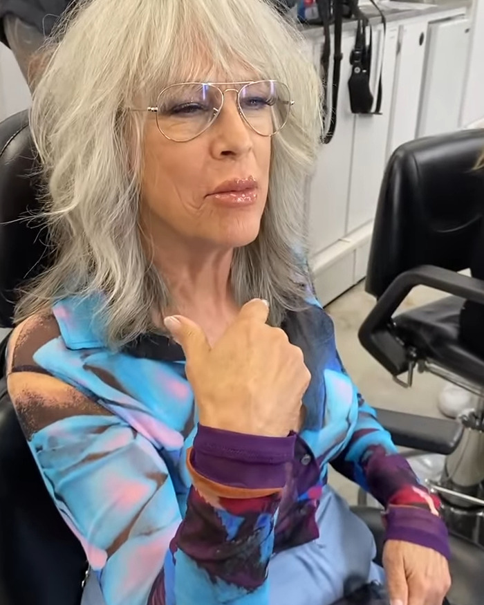 Jamie Lee Curtis with dramatically fuller lips, wearing glasses and a colorful shirt, sitting in a salon chair. Jamie Lee Curtis with dramatically fuller lips, wearing glasses and a colorful shirt, sitting in a salon chair.