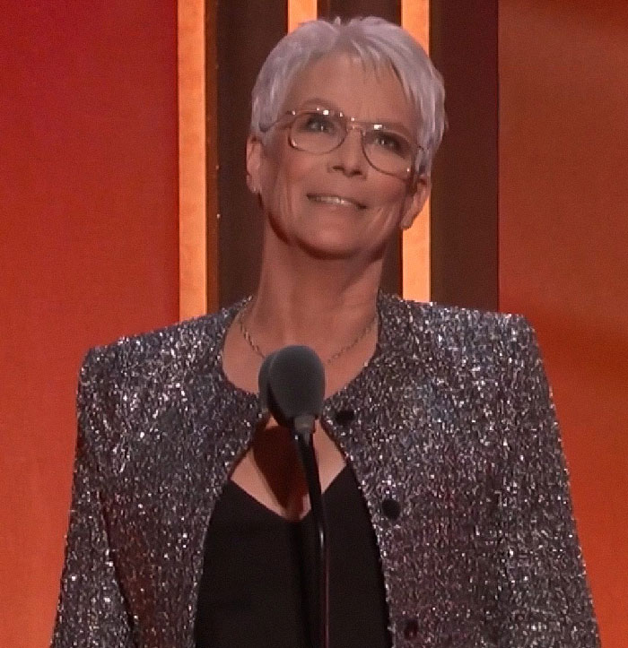 Jamie Lee Curtis speaking at an event wearing glasses and a sparkly jacket with a microphone in front of her.