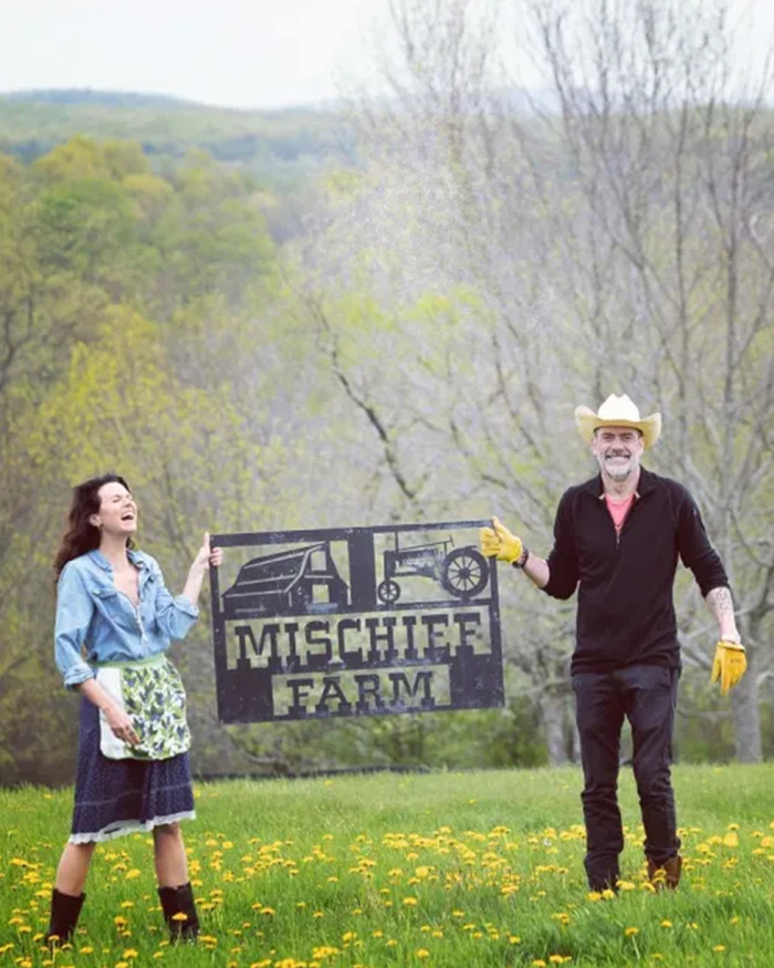 Jeffrey Dean Morgan and a woman holding a Mischief Farm sign outdoors in a field with trees in the background.