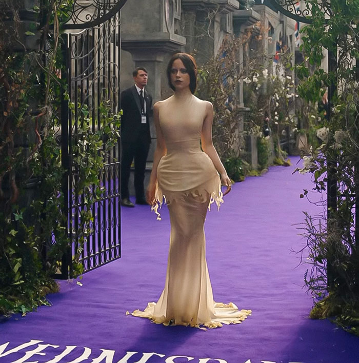 Jenna Ortega wearing a sleek beige gown on a purple carpet at an event, showcasing Wednesday looks in fashion.