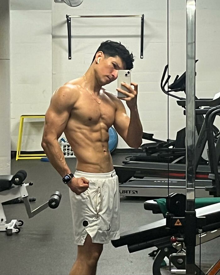 Muscular man in white shorts taking a gym mirror selfie, showcasing his toned bikini body and fit physique.