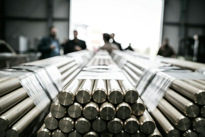 Stack of shiny metal rods in a warehouse setting with blurred workers in the background, illustrating new facts and learning concepts.