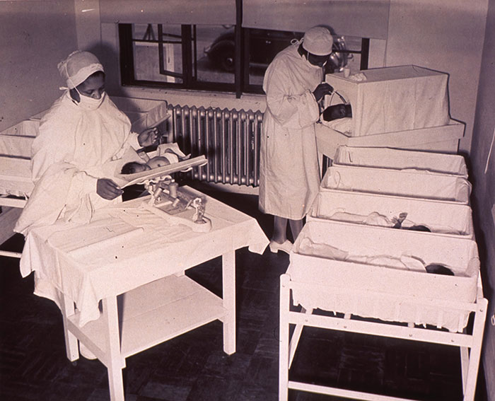 Two nurses in vintage uniforms caring for infants in cribs in a dimly lit hospital nursery room.