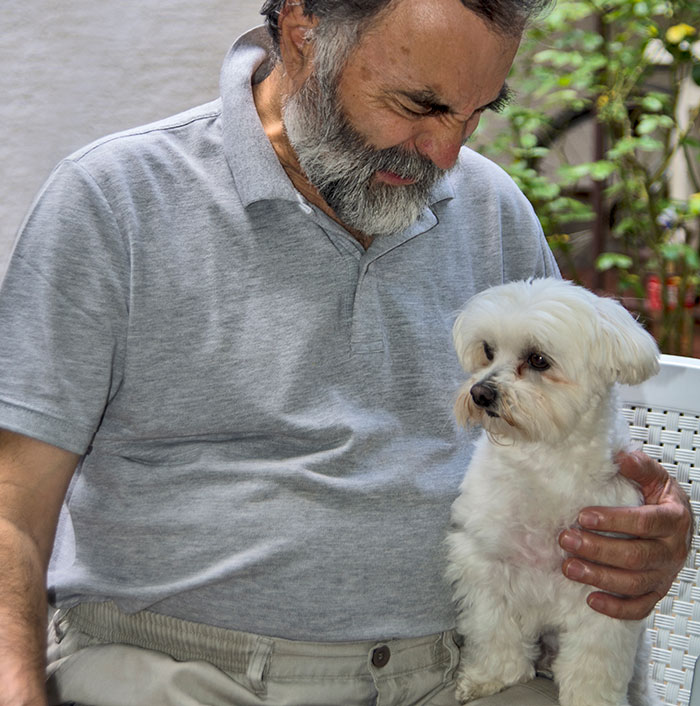Man with gray beard sitting and comforting a small white dog, illustrating survival and recovery from kidnapping experiences.