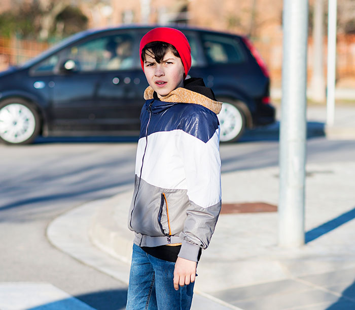 Teen boy wearing a red beanie and jacket standing on a street near a parked black car, appearing alert and cautious