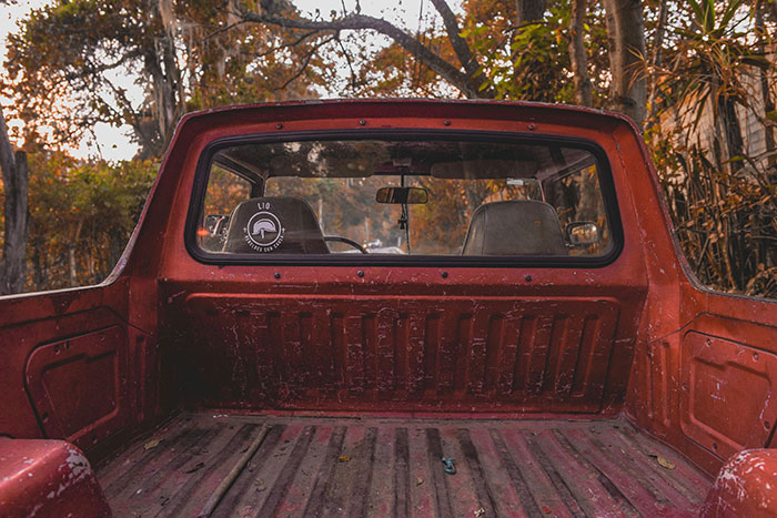 Red rusty pickup truck bed with seats visible through the rear window in a wooded autumn setting, evoking kidnapped survivor stories.