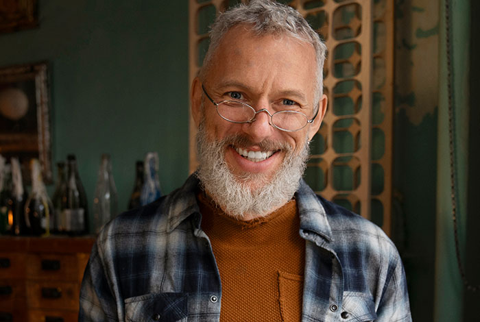 Smiling older man with glasses and gray beard sharing surviving kidnapping experiences in a cozy indoor setting.