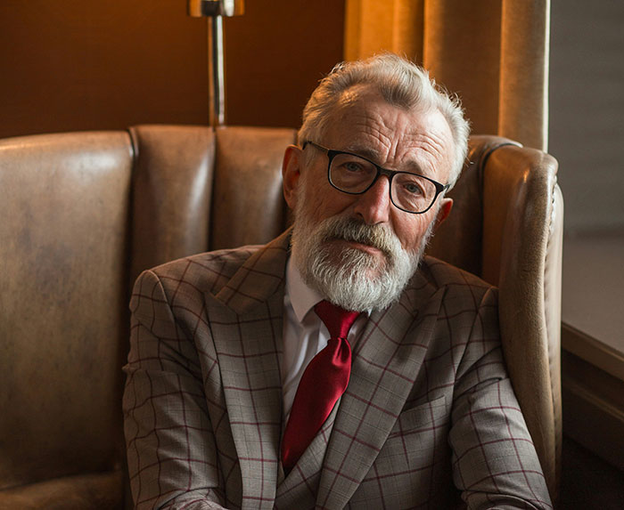 Elderly man with glasses and beard sitting in leather chair reflecting on kidnapping survivor experiences.